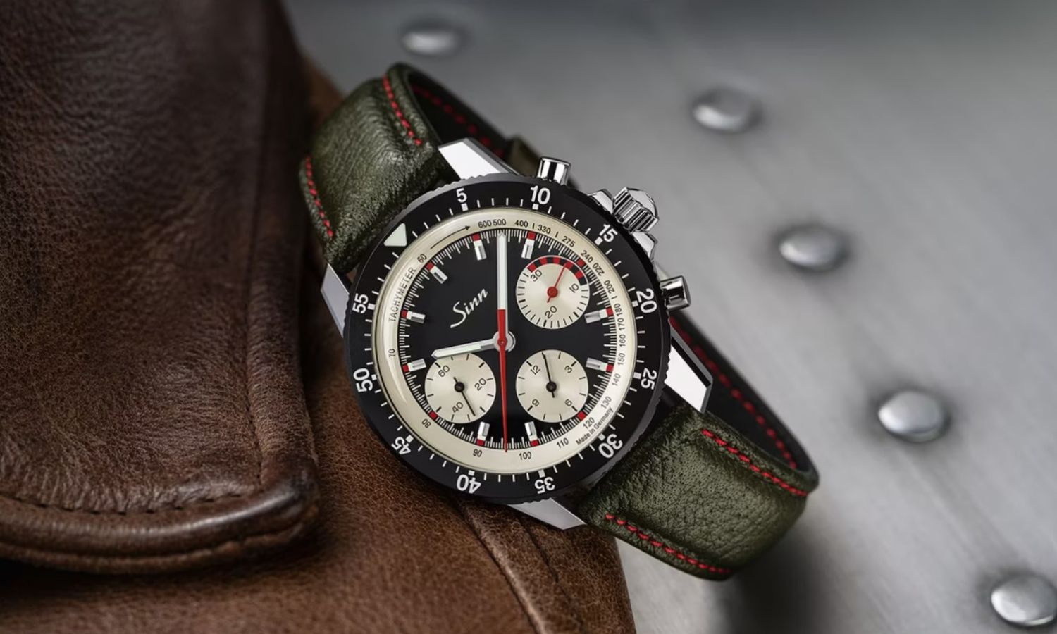 Sinn 103 ST Ty Hd Limitierte Edition mit schwarzem Zifferblatt und grünem Armbanfdf liegend auf einem Lederprodukt vor grauem Industrial-Hintergrund