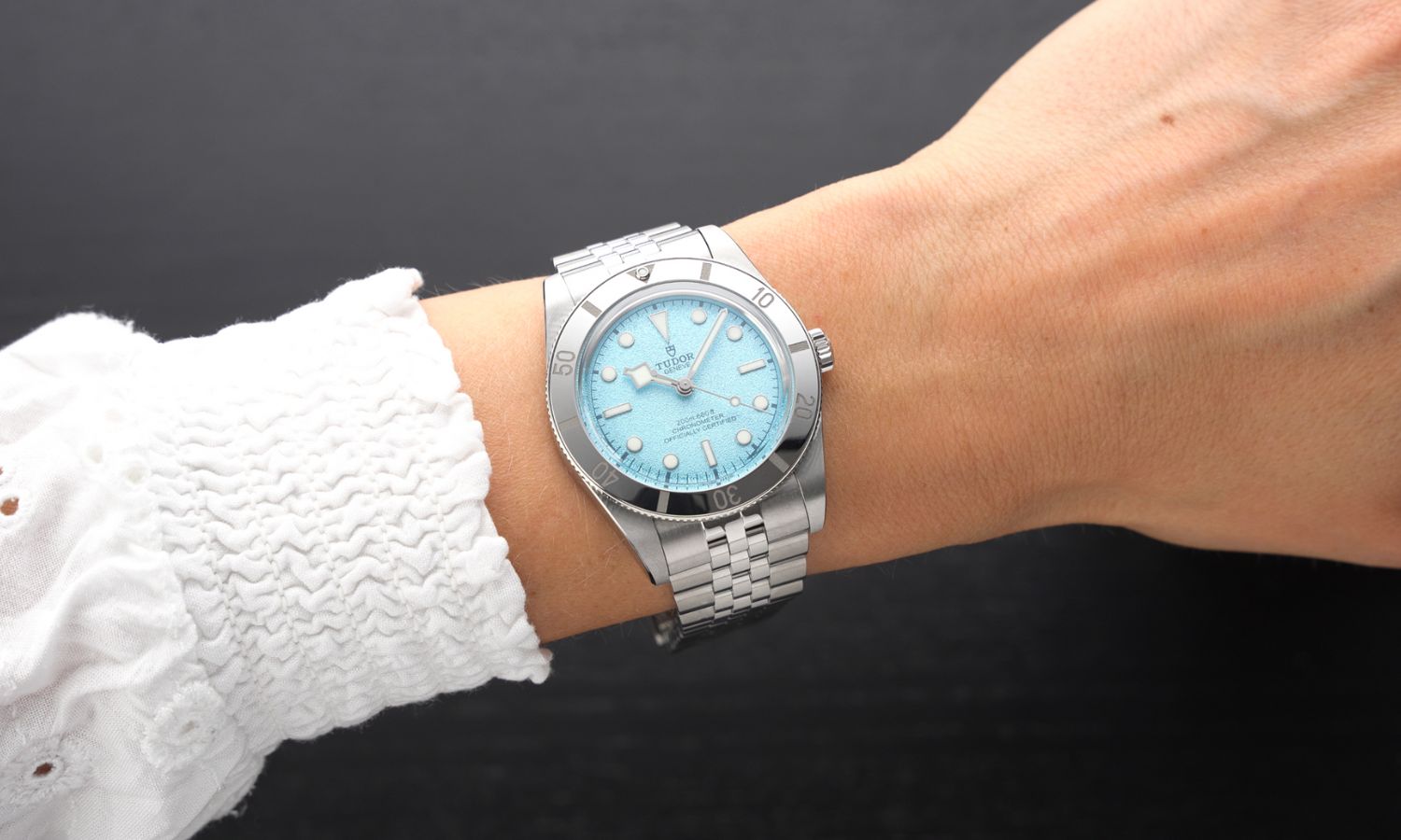 TUDOR Black Bay 54 Lagoon Blue Referenz: M79000-0001 mit hellblauem Zifferblatt am Edelstahlarmband an einem weiblichen Handgelenk mit weißem Ärmel getragen vor dunkelgrauem Hintergrund