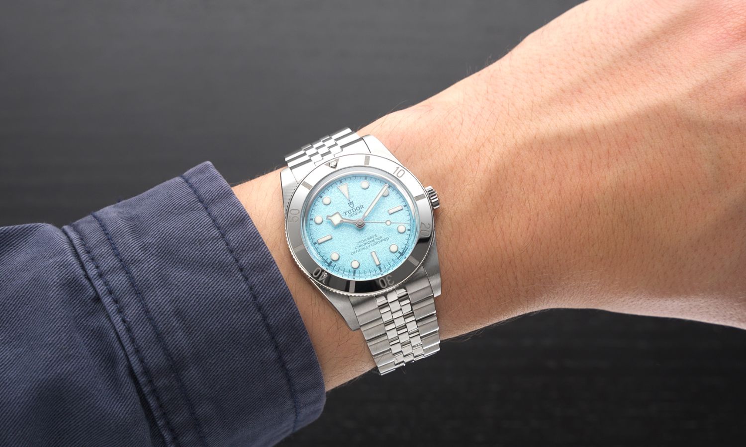 TUDOR Black Bay 54 Lagoon Blue Referenz: M79000-0001 mit hellblauem Zifferblatt am Edelstahlarmband an einem männlichen Handgelenk mit dunkelblauem Ärmel getragen vor dunkelgrauem Hintergrund