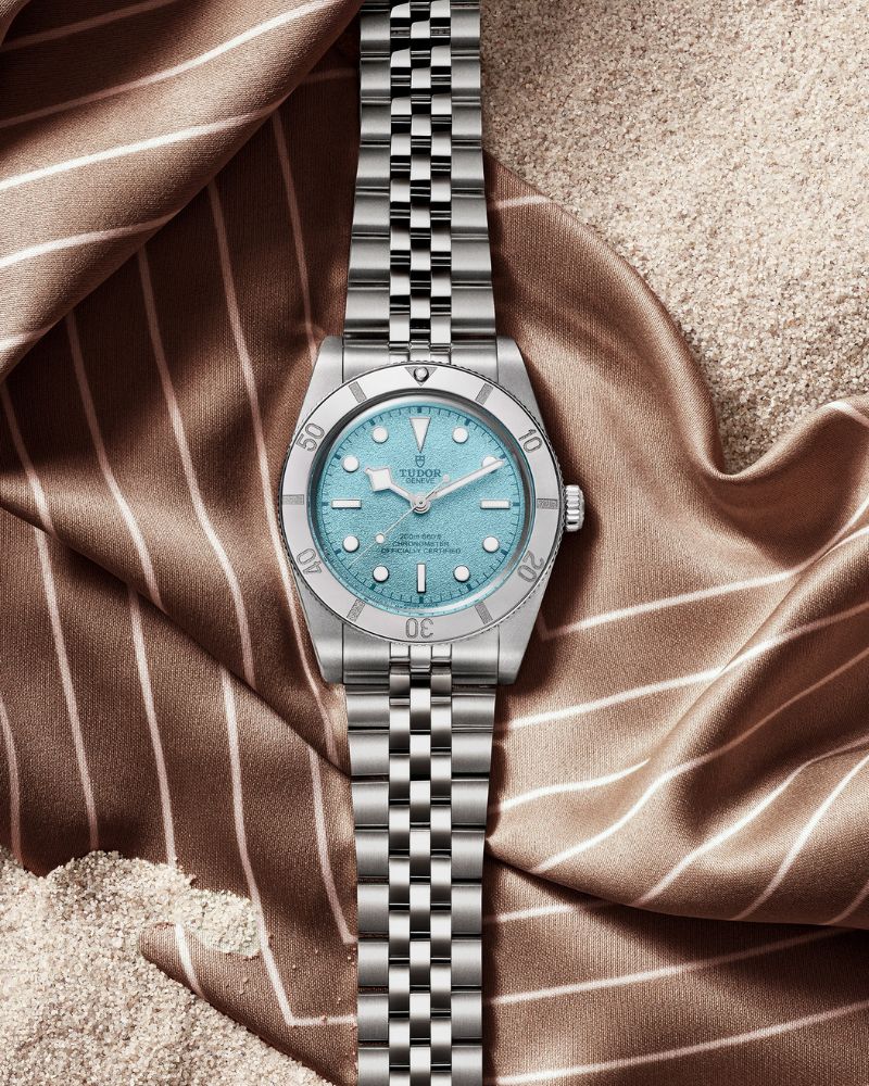 Blick von oben auf die TUDOR Black Bay 54 Lagoon Blue mit der Referenz 79000 und blauem Zifferblatt, die auf einem Tuch auf einem Sandstrand liegt.