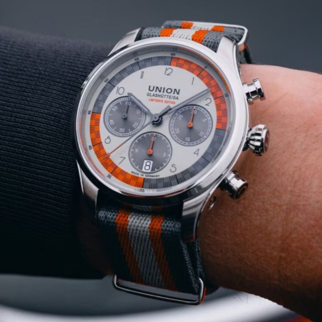 Union Glashütte Belisar Chronograph Silvretta Classic 2025 am grau-orangenen Natoband abgebildet an einem Handgelenk mit schwarzem Ärmel vor dunklem Hintergrund