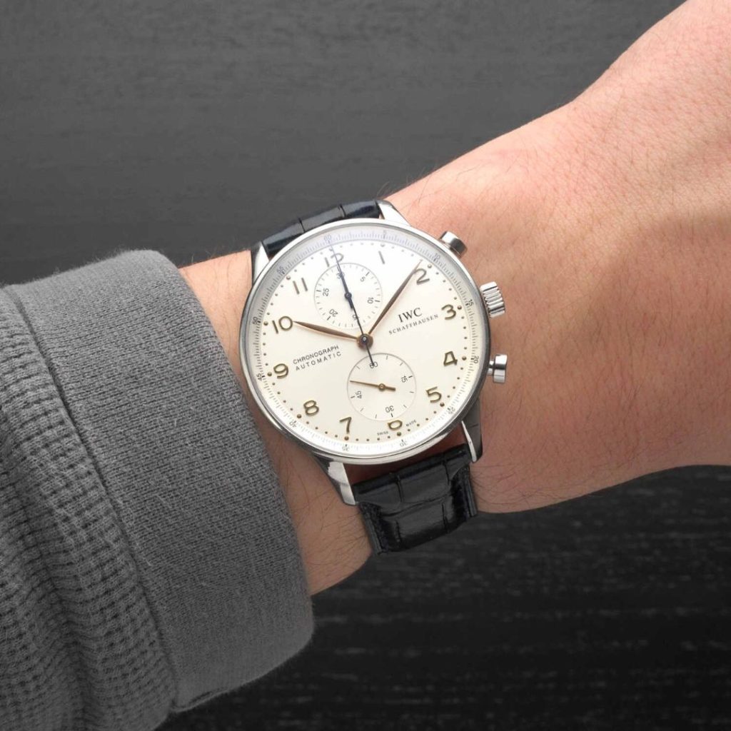 IWC Schaffhausen Portugieser Chronograph mit weißem Zifferblatt am schwarzen Lederarmband an einem Handgelenk mit grauem Ärmel getragen vor dunkelgrauem Hintergrund