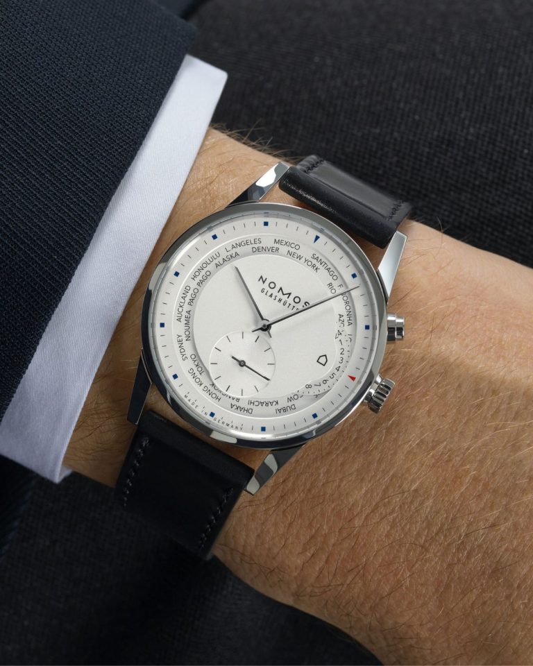NOMOS Glashütte Zürich Weltzeit Referenz 805 mit weißem Zifferblatt und schwarzem Lederarmband abgebildet an einem Handgelenk mit dunklem Jackett und weißem Hemd vor dunklem Hintergrund