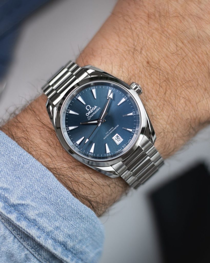 Omega Seamaster Aqua Terra 38 mit blauem Zifferblatt am Edelstahlarmband abgebildet an einem Handgelenk mit hellblauem Ärmel vor weißem Hintergrund mit Schatten