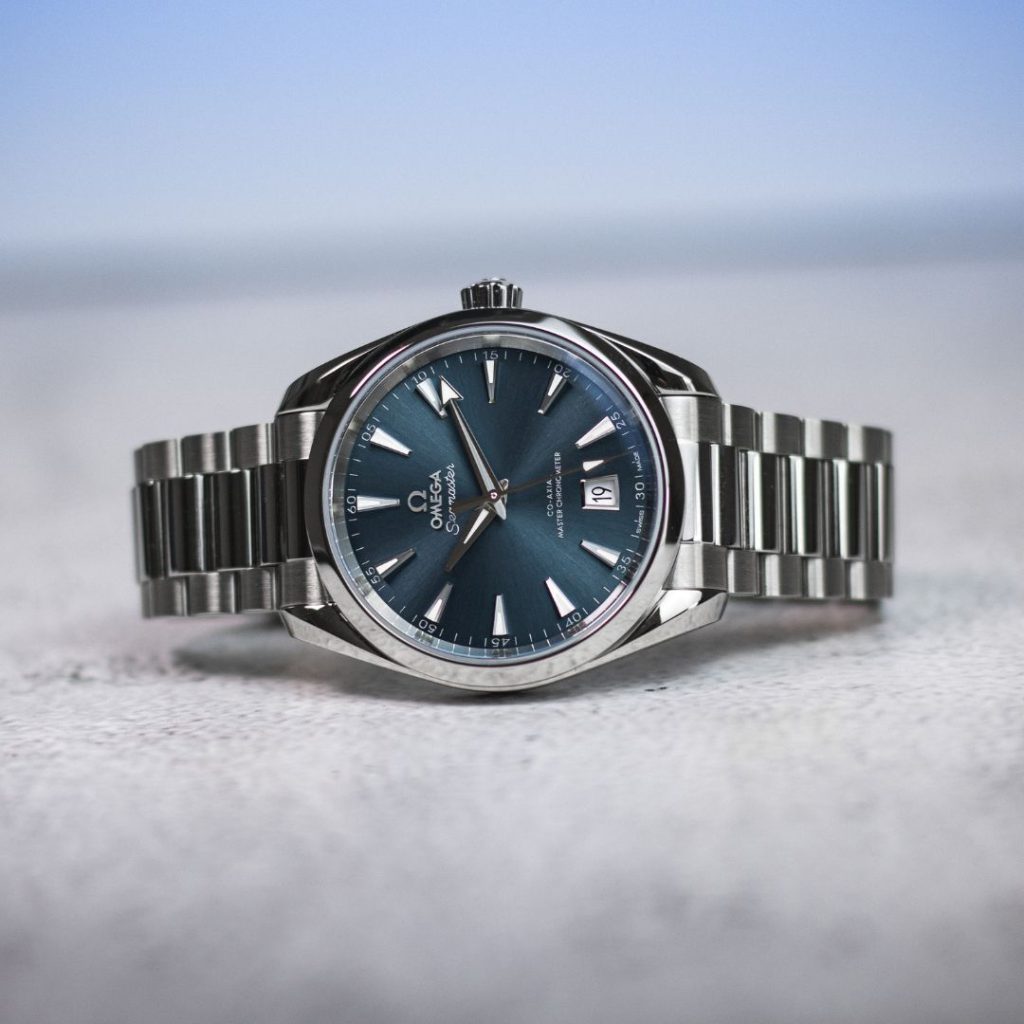 OMEGA Seamaster Aqua Terra 38 mit blauem Zifferblatt am Edelstahlarmband abgebildet auf einem weißen Untergrund vor hellblauem Hintergrund
