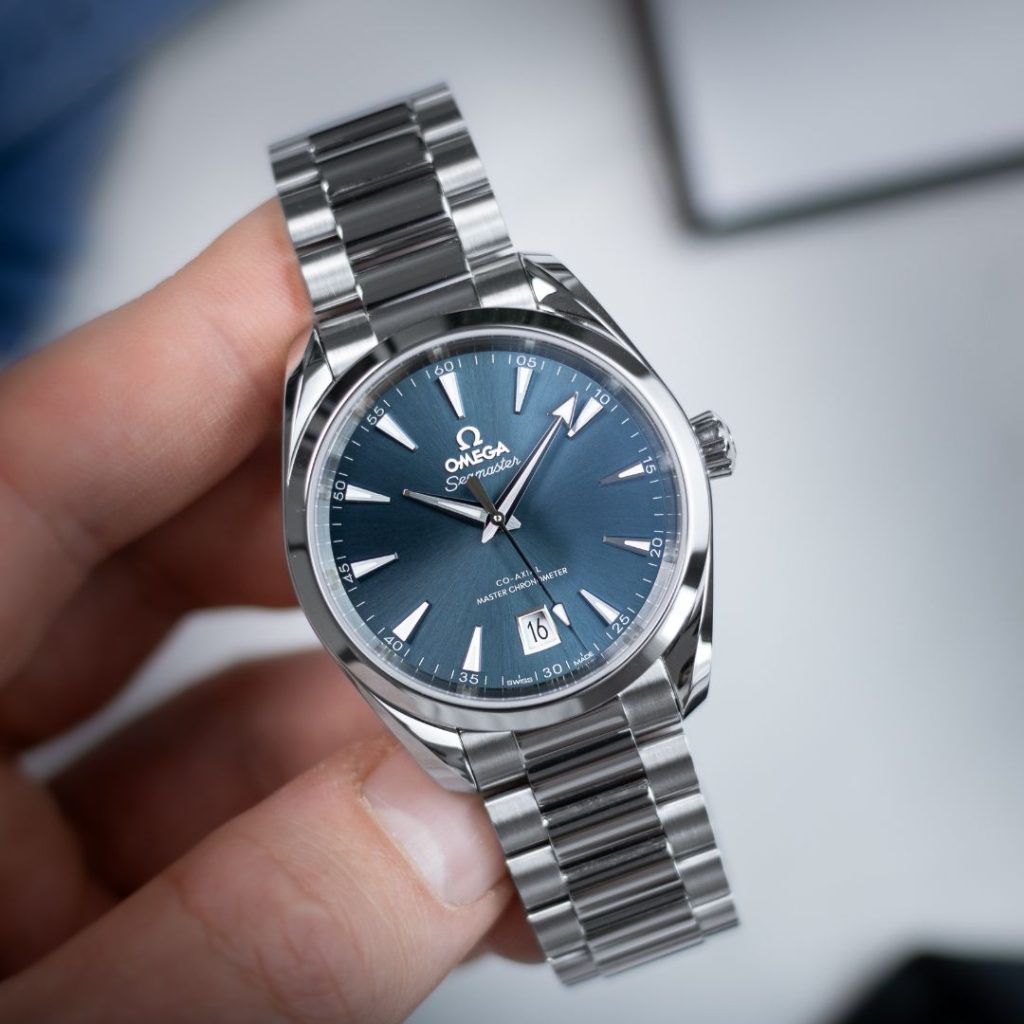 OMEGA Seamaster Aqua Terra 38 mit blauem Zifferblatt und Edelstahlarmband gehalten von einer Hand vor weißem Hintergrund mit verschwommenen Gegenständen