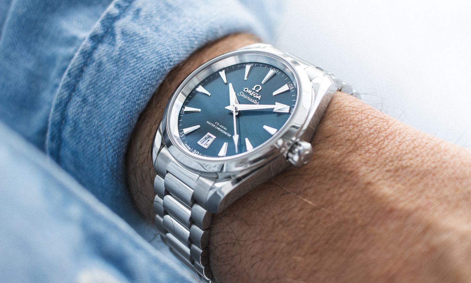 OMEGA Seamaster Aqua Terra 38 mit blauem Zifferblatt und Edelstahlarmband abgebildet an einem Handgelenk mit blauem Ärmel vor weißem Hintergrund