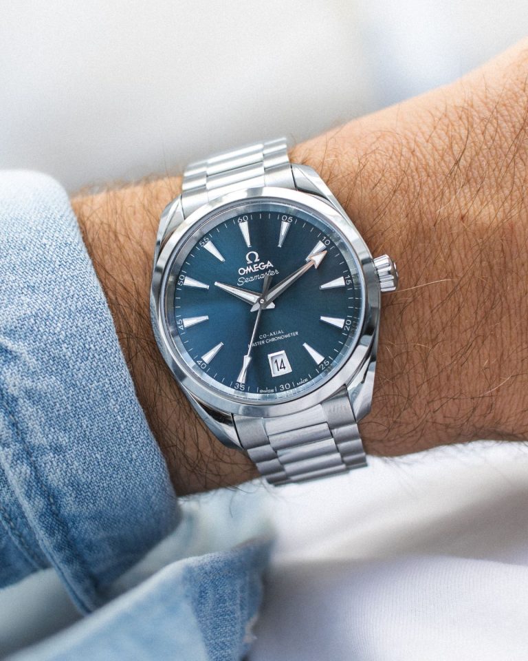 Omega Seamaster Aqua Terra 38 mit blauem Zifferblatt am Edelstahlarmband abgebildet an einem Handgelenk mit hellblauem Ärmel vor weißem Hintergrund