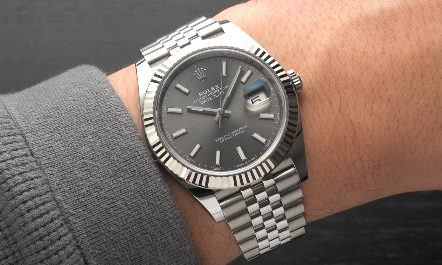 Rolex Datejust 41 Referenz 126334 mit grauem Zifferblatt am Jubilé Armband an einem Handgelenk mit grauem Ärmel getragen vor grauem Hintergrund