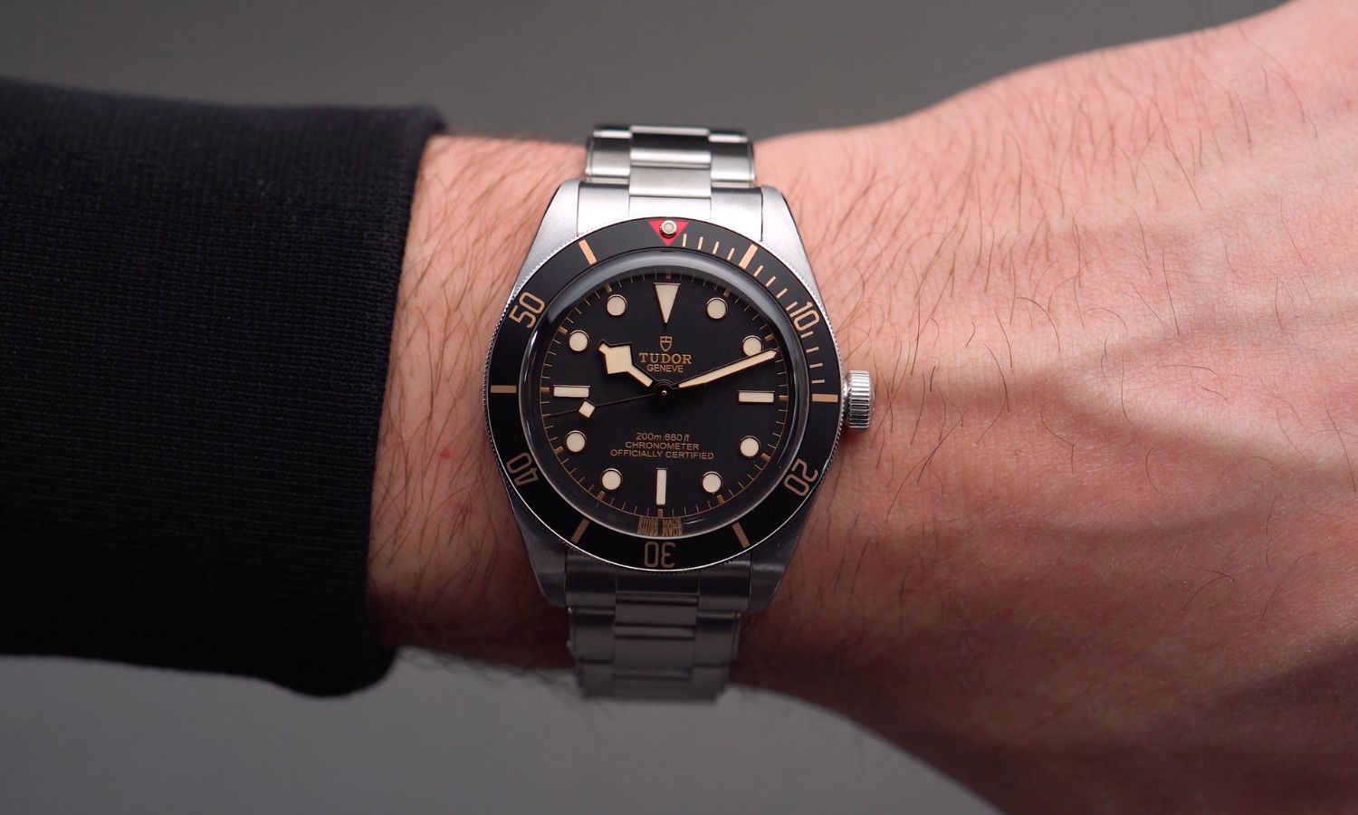 TUDOR Black Bay 58 Referenz M79030N-0001 mit schwarzem Zifferblatt und schwarzer Lünette am Edelstahlarmband an einem Handgelenk mit schwarzem Ärmel getragen vor grauem Hintergrund