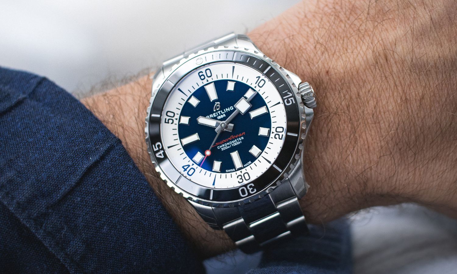 Breitling Superocean Automatic 44 mit blauem Zifferblatt am Edelstahlarmband an einem Handgelenk mit dunkelblauem Ärmel getragen vor grauem Hintergrund