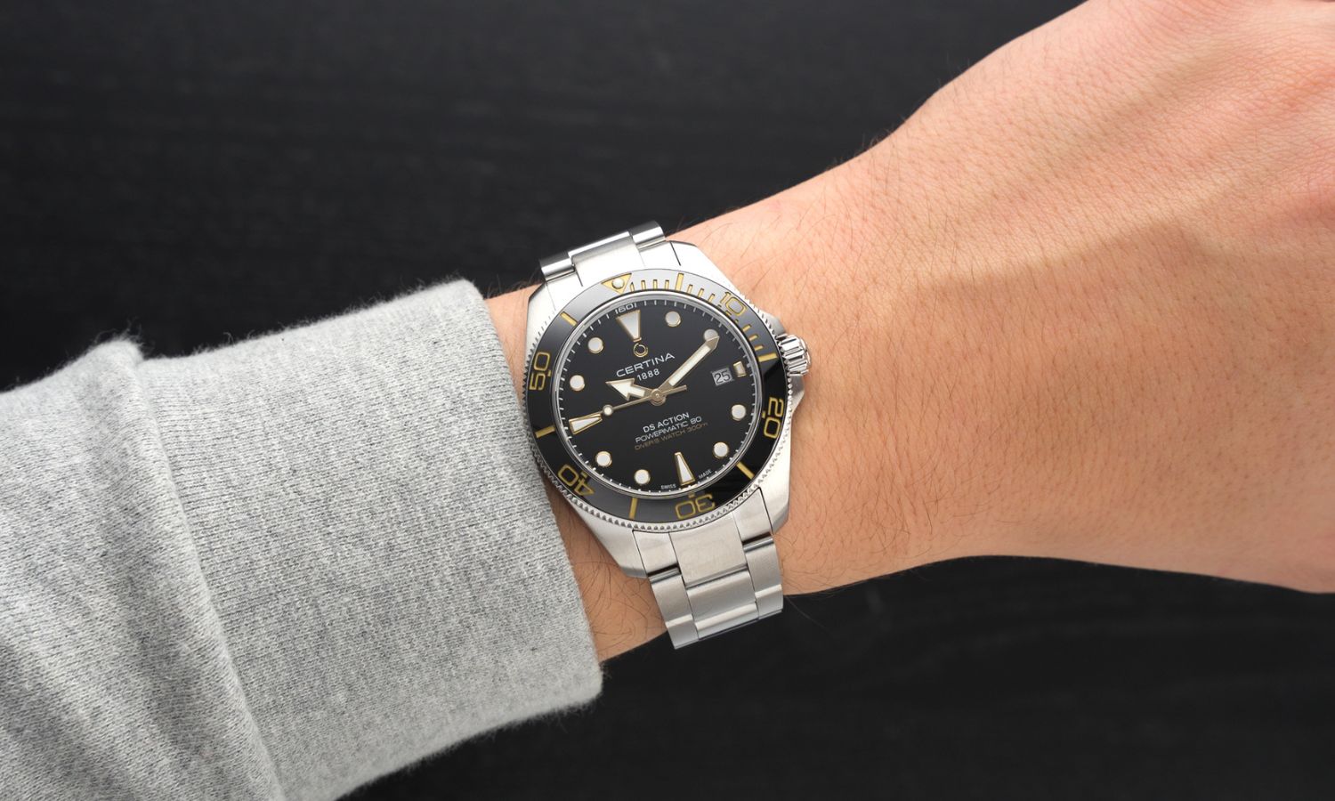 Certina DS Action Diver 40,5mm Powermatic 80 Referenz: C048.407.11.051.01 mit schwarzem Zifferblatt am Edelstahlarmband an einem Handgelenk mit hellgrauem Ärmel getragen vor schwarzem Hintergrund
