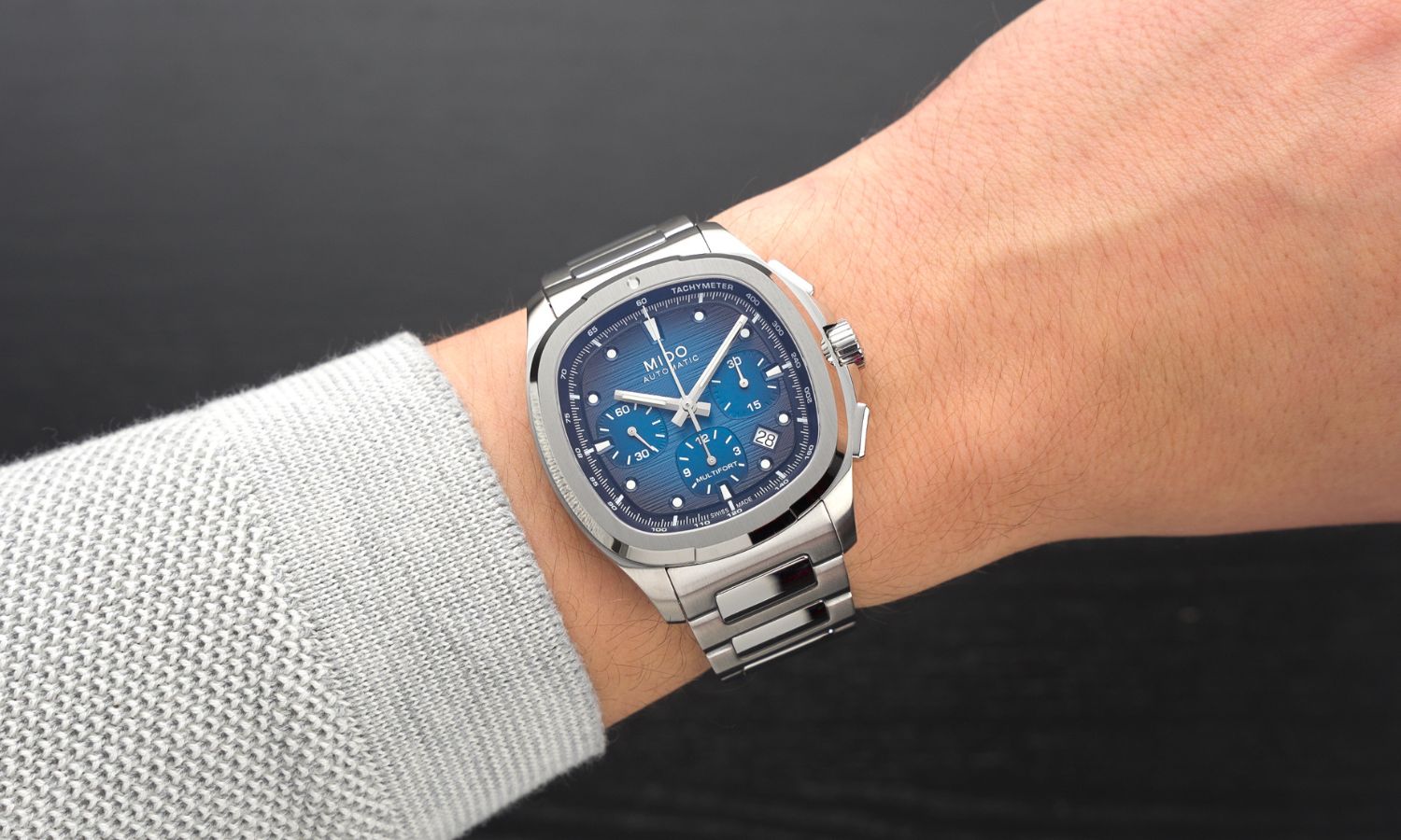 Mido Multifort TV Chronograph Referenz: M049.527.11.041.00 mit blauem Zifferblatt am Edelstahlarmband an einem Handgelenk mit grauem Ärmel getragen vor schwarzem Hintergrund