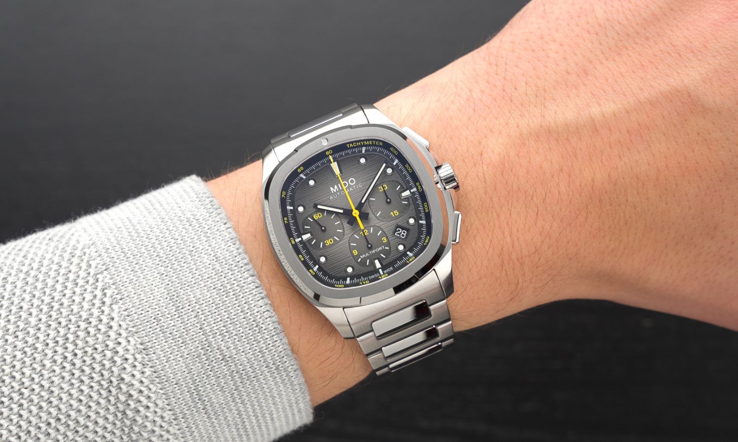 Mido Multifort TV Chronograph Referenz: M049.527.11.081.00 mit grauem Zifferblatt am Edelstahlarmband an einem Handgelenk mit grauem Ärmel getragen vor schwarzem Hintergrund
