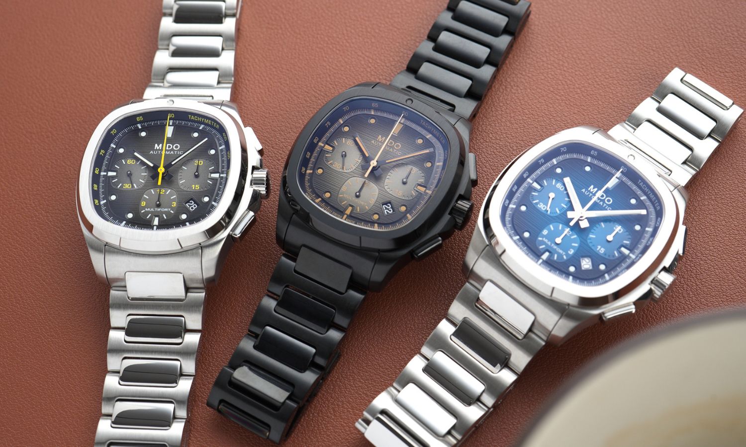 Drei Modelle des Mido Multifort TV Chronographen nebeneinander liegend vor braunem Hintergrund