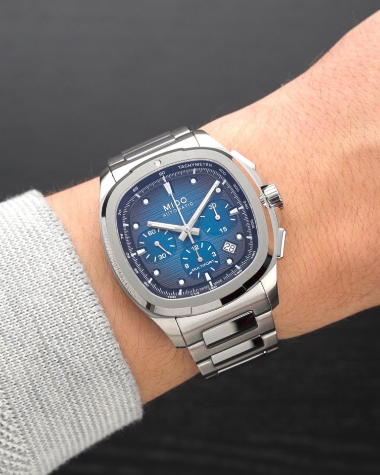 Mido Multifort TV Chronograph Referenz: M049.527.11.041.00 mit blauem Zifferblatt am Edelstahlarmband an einem Handgelenk mit grauem Ärmel getragen vor schwarzem Hintergrund