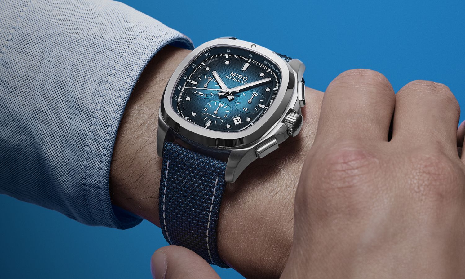 Mido Multifort TV Chronograph Referenz: M049.527.11.041.00 mit blauem Zifferblatt am blauen Synthetikarmband an einem Handgelenk mit grauem Ärmel getragen vor blauem Hintergrund