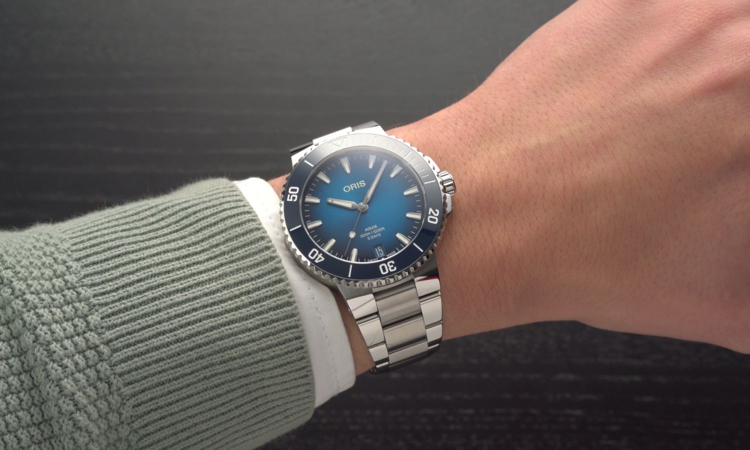 Oris Aquis Date Calibre 400 mit blauem Zifferblatt am Edelstahlarmband an einem Handgelenk mit grünem Ärmel getragen vor dunkelgrauem Hintergrund