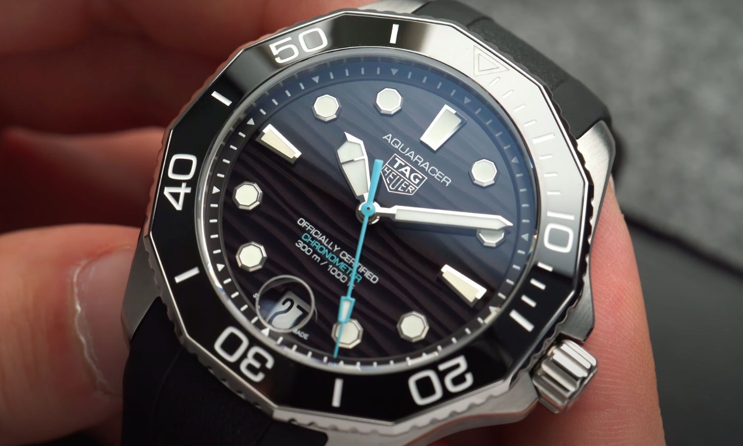 Nahaufnahme der TAG HEUER Aquaracer Professional 300 Date mit schwarzem Zifferblatt am schwarzen Kautschukarmband in zwei Händen gehalten vor grauem Hintergrund