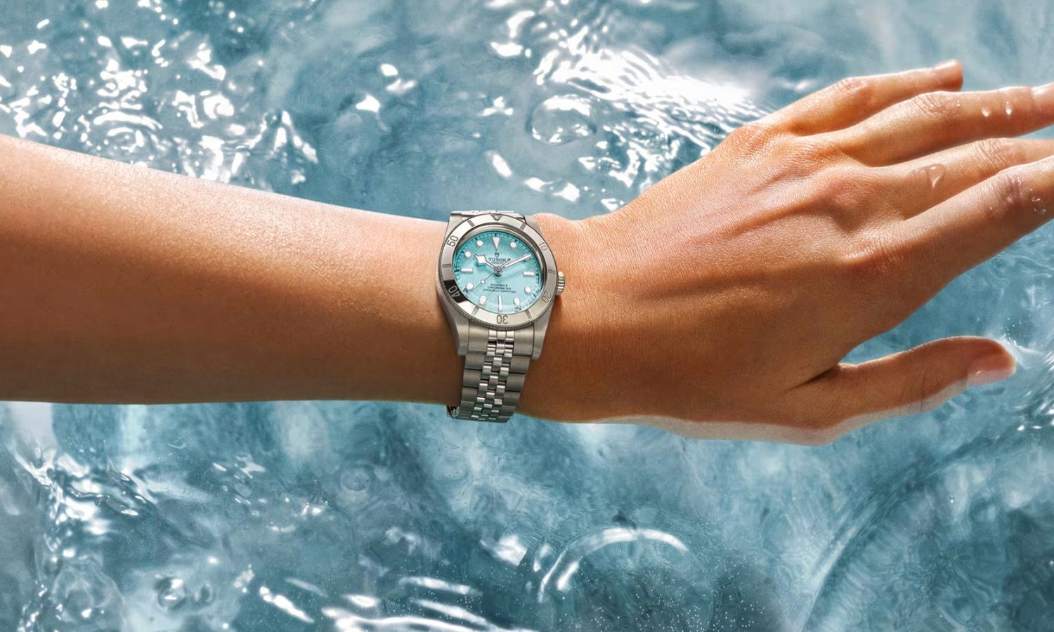TUDOR Black Bay 54 Lagoon Blue mit hellblauem Zifferblatt am Edelstahlarmband an dem Handgelenk einer Frau getragen mit Wasser im Hintergrund
