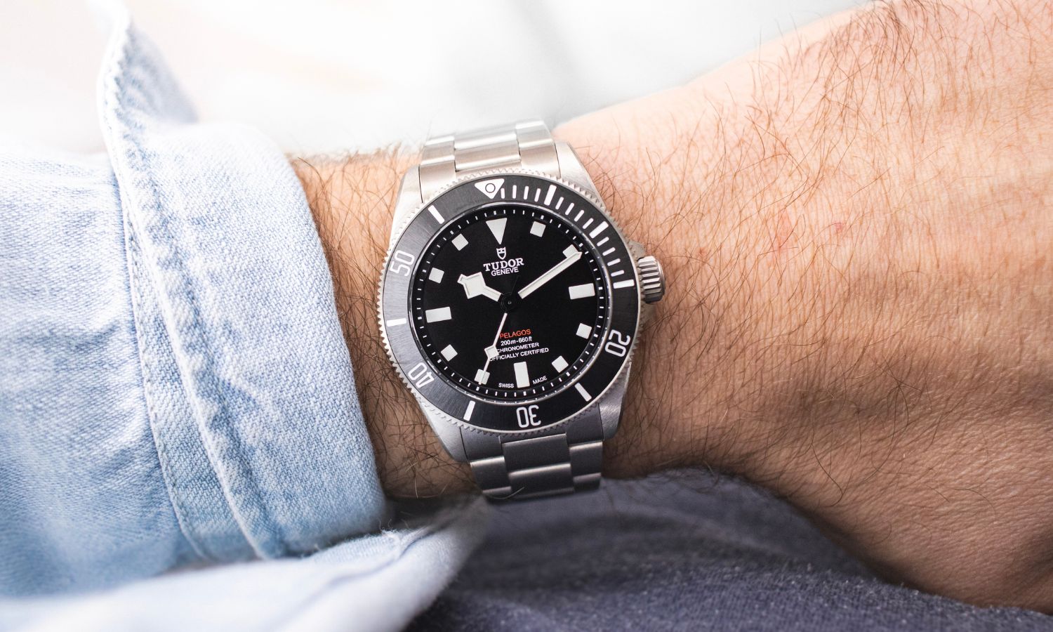 TUDOR Pelagos 39 mit schwarzem Zifferblatt am Edelstahlarmband an einem Handgelenk mit hellblauem Ärmel getragen vor hellem Hintergrund