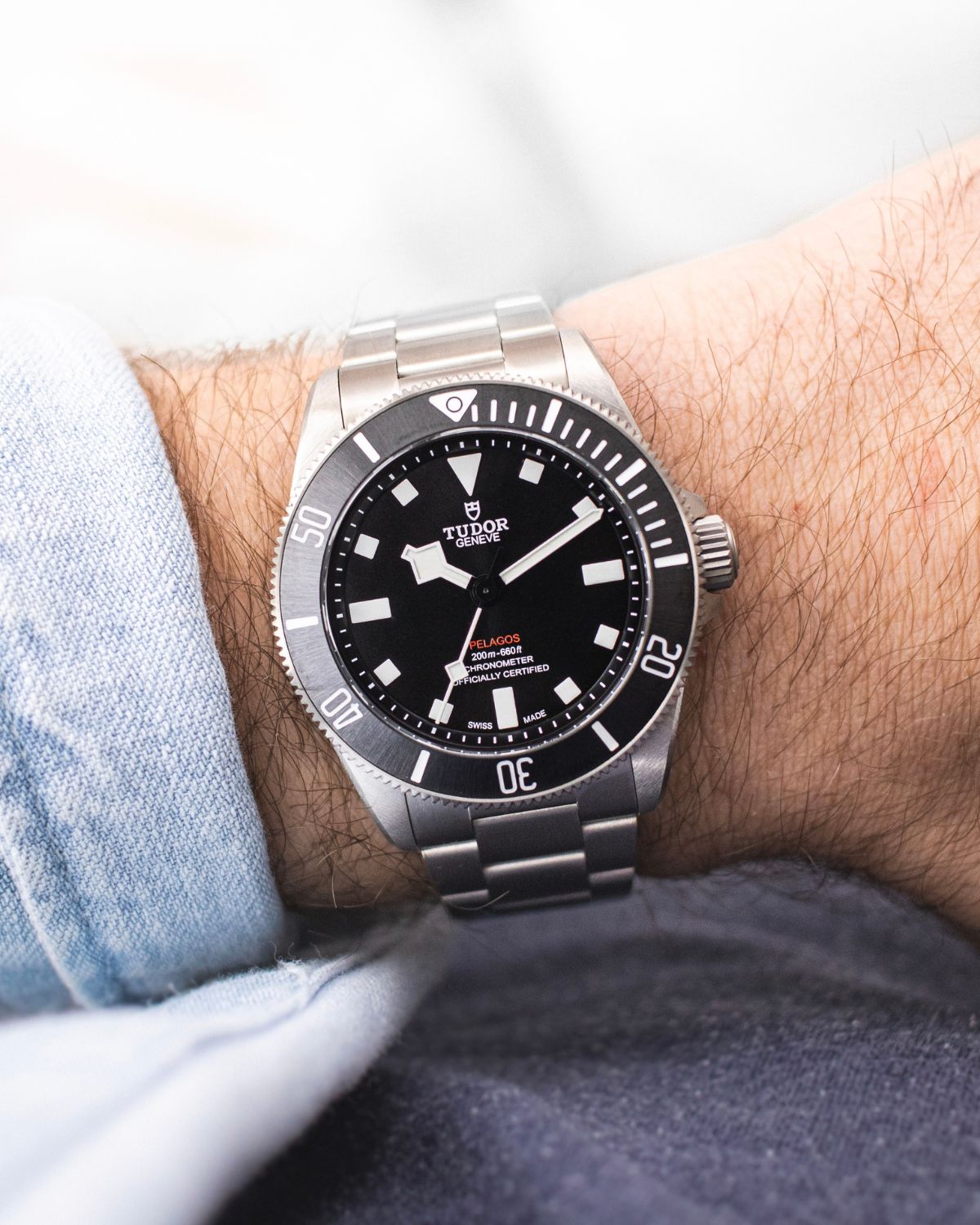 TUDOR Pelagos 39 mit schwarzem Zifferblatt am Edelstahlarmband an einem Handgelenk mit hellblauem Ärmel getragen vor hellem Hintergrund