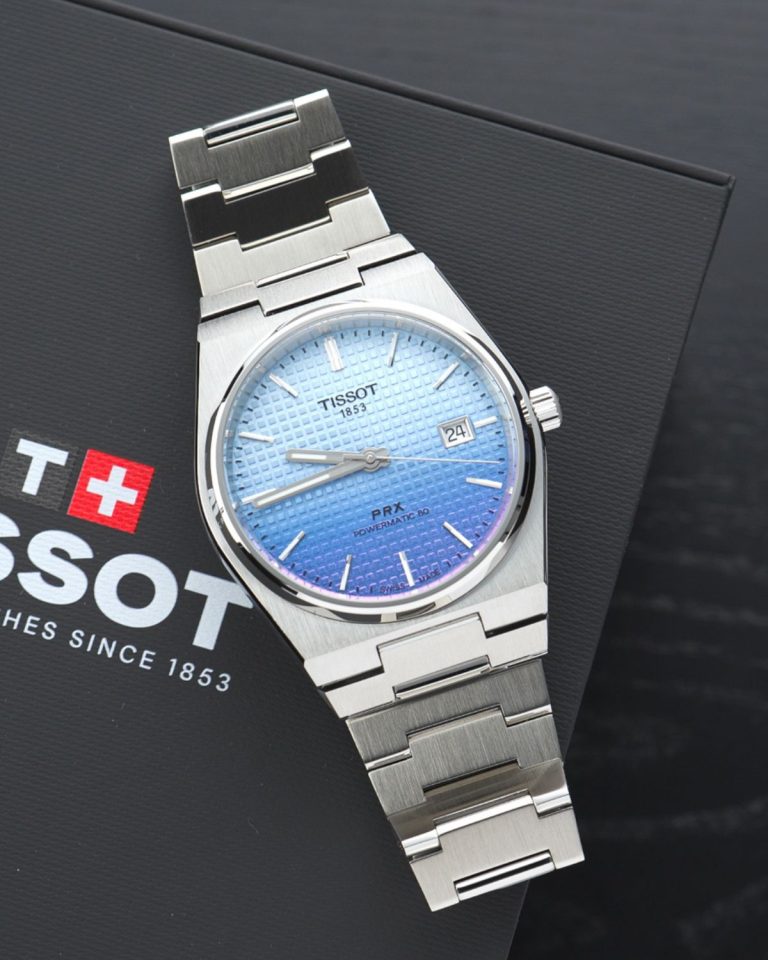 Tissot PRX Powermatic 80 Gradient Referenz T137.407.11.351.01 mit blau-violettem Zifferblatt am Edelstahlarmband liegend auf einer schwarzen Tissot Box vor schwarzem Hintergrund