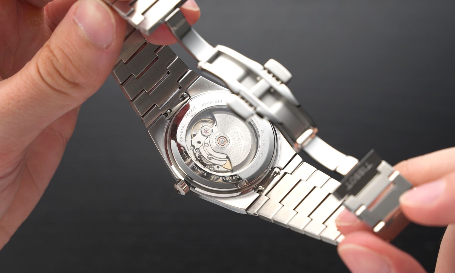 Die Tissot PRX Powermatic 80 Gradient Referenz T137.407.11.351.01 in zwei Händen gehalten mit Blick auf das Powermatic 80 Uhrwerk sowie auf das Edelstahlarmband und die geöffnete Schließe der Uhr vor schwarzem Hintergrund