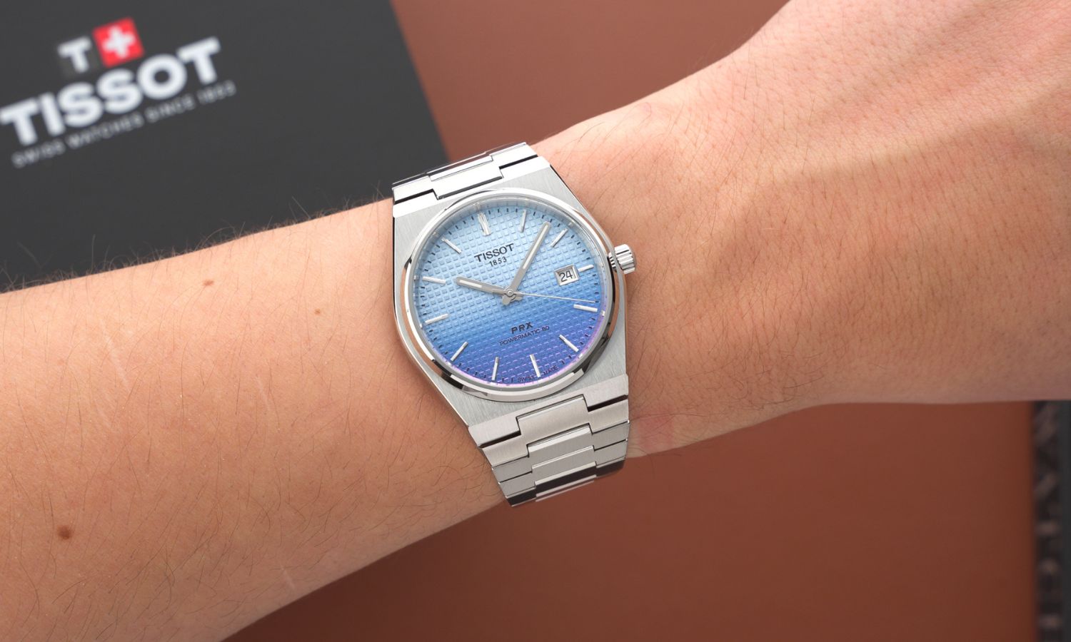 Tissot PRX Powermatic 80 Gradient Referenz T137.407.11.351.01 mit blau-violettem Zifferblatt am Edelstahlarmband an einem Handgelenk getragen vor einer schwarzen Tissot Box und hellbraunem Hintergrund