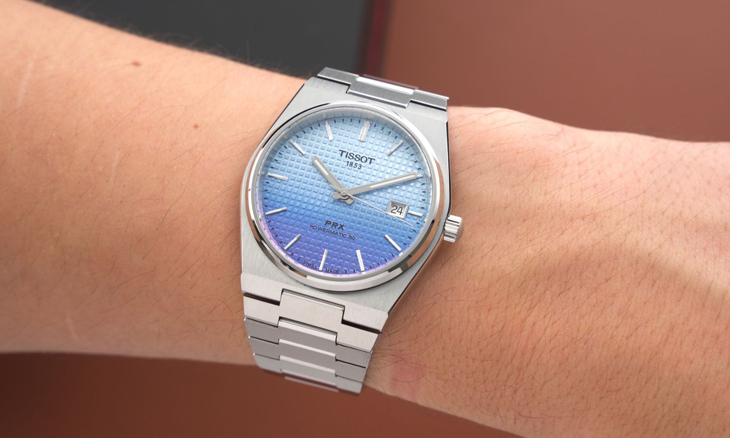 Tissot PRX Powermatic 80 Gradient Referenz T137.407.11.351.01 mit blau-violettem Zifferblatt am Edelstahlarmband an einem Handgelenk getragen vor einer schwarzen Tissot Box und hellbraunem Hintergrund