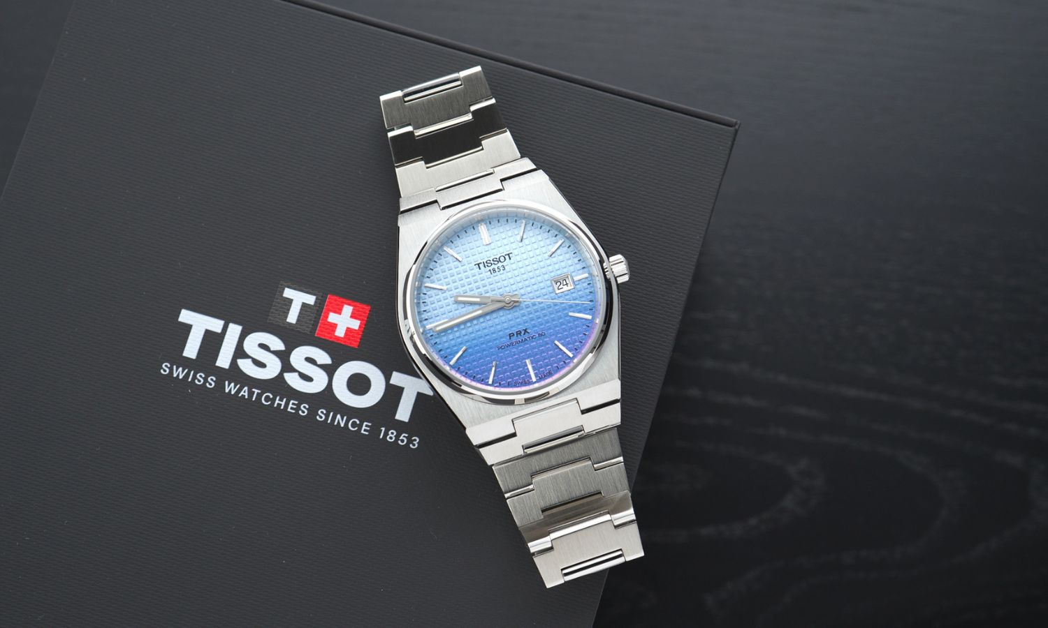 Tissot PRX Powermatic 80 Gradient Referenz T137.407.11.351.01 mit blau-violettem Zifferblatt am Edelstahlarmband liegend auf einer schwarzen Tissot Box vor schwarzem Hintergrund