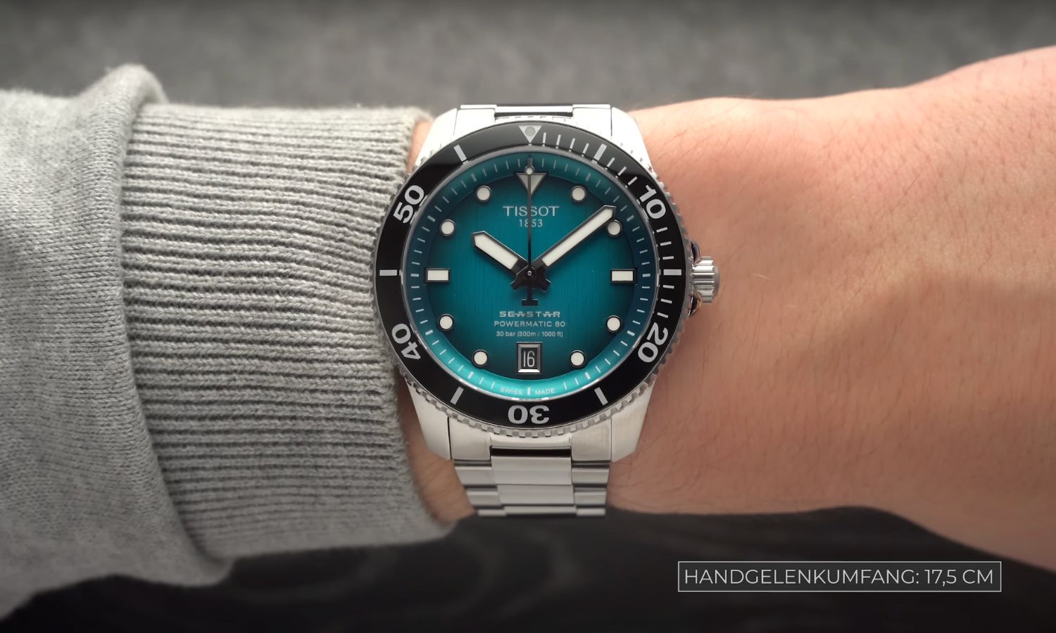 Tissot Seastar 1000 Powermatic 80 40 mm mit türkisem Zifferblatt am Edelstahlarmband an einem Handgelenk mit hellgrauem Ärmel getragen vor grauem Hintergrund