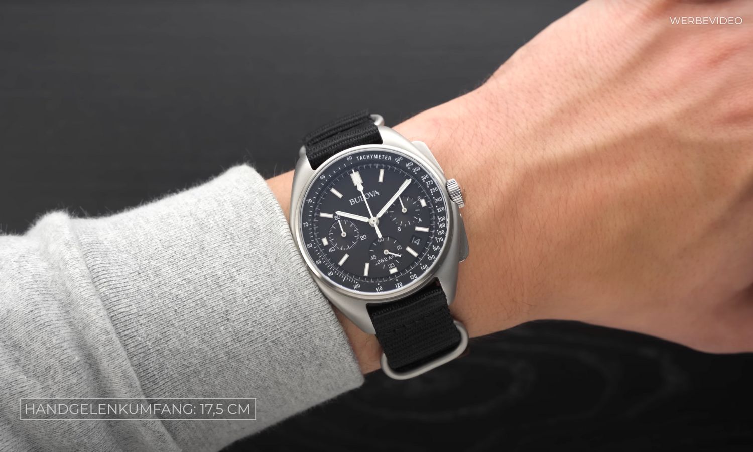 Bulova Lunar Pilot mit schwarzem Zifferblatt am schwarzen NATO-Band an einem Handgelenk mit grauem Ärmel getragen vor schwarzem Hintergrund