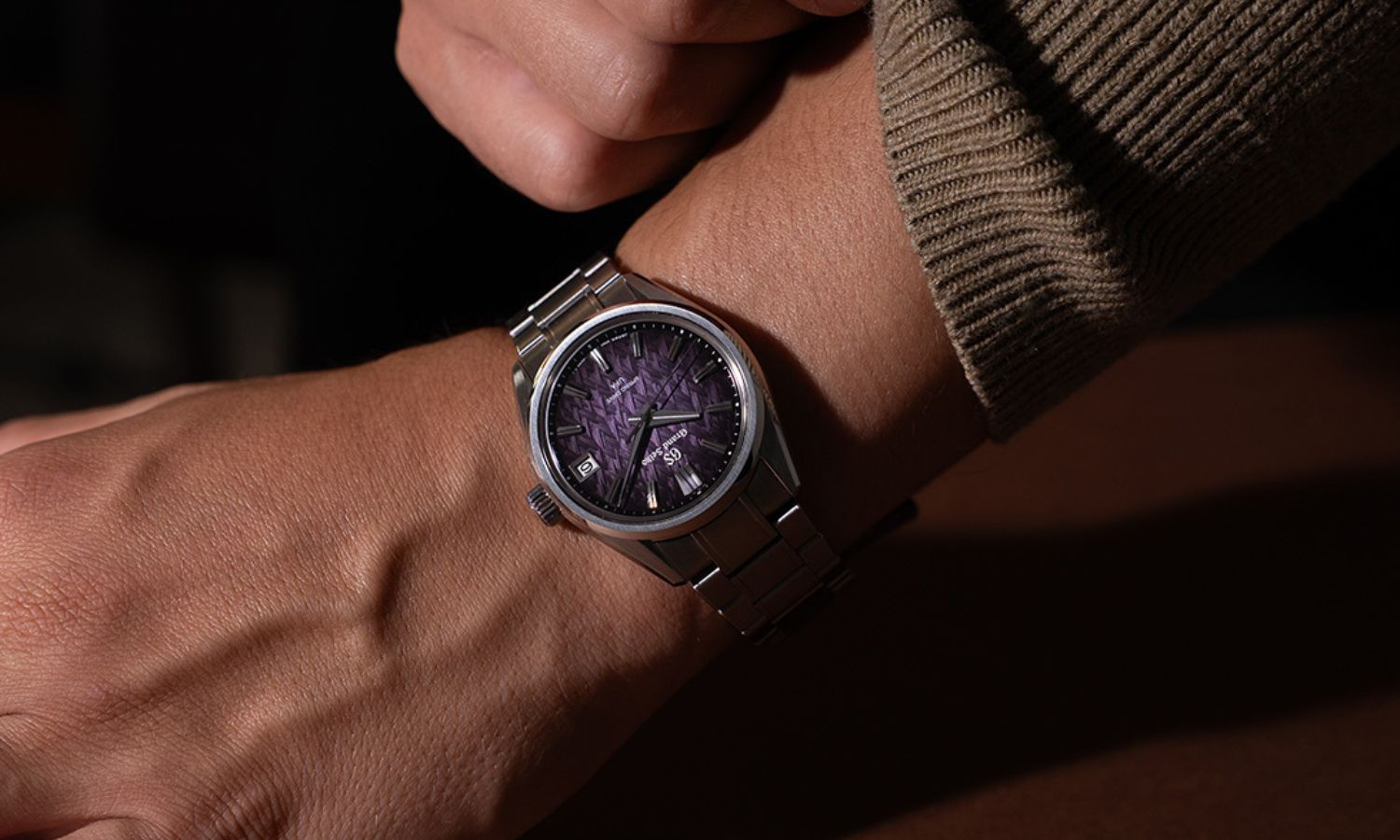 Grand Seiko SLGB005 „Violet Dawn“ mit violettem Zifferblatt am Edelstahlarmband an einem Handgelenk mit braunem Ärmel getragen vor dunklem Hintergrund