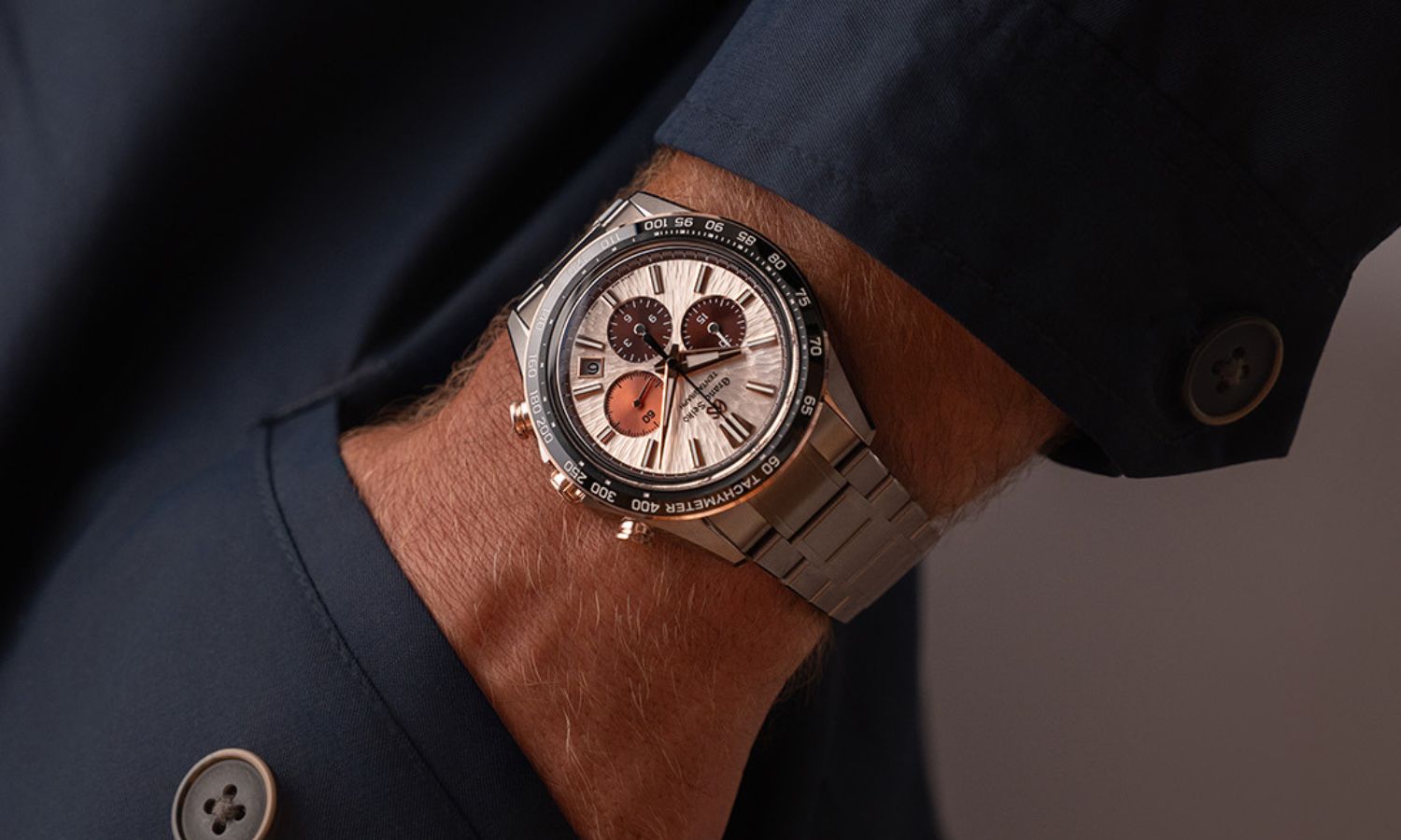 Grand Seiko SLGC006 Sunrise Tentagraph mit kupferfarbenem Zifferblatt am Edelstahlarmband an einem Handgelenk mit dunkelblauem Ärmel mit der Hand in der Hosentasche getragen