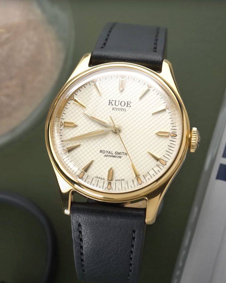 Kuoe Royal Smith Gold Ivory mit cremefarbenem Zifferblatt am schwarzen Lederarmband stehend vor dunkelgrünem Hintergrund