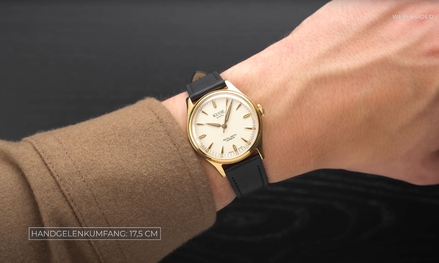 Kuoe Royal Smith Gold Ivory mit cremefarbenem Zifferblatt am schwarzen Lederarmband an einem Handgelenk mit hellbraunem Ärmel getragen vor schwarzem Hintergrund