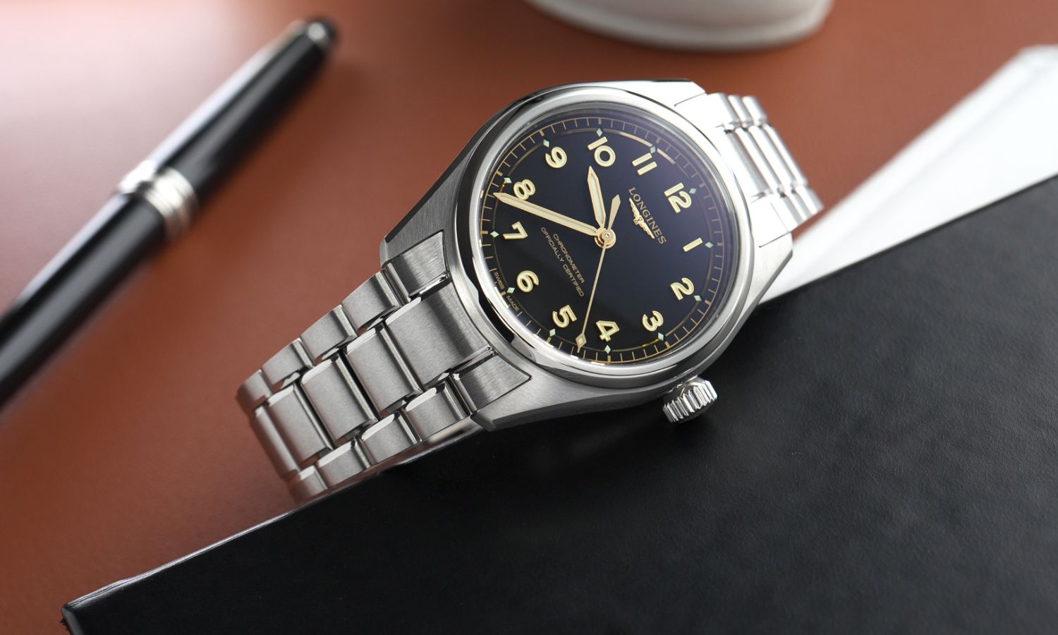 Longines Spirit Pilot Referenz L3.809.4.53.6 mit schwarzem Zifferblatt am Edelstahlarmband von vorne abgebildet seitlich liegend neben einem schwarzen Kugelschreiber vor braunem Hintergrund