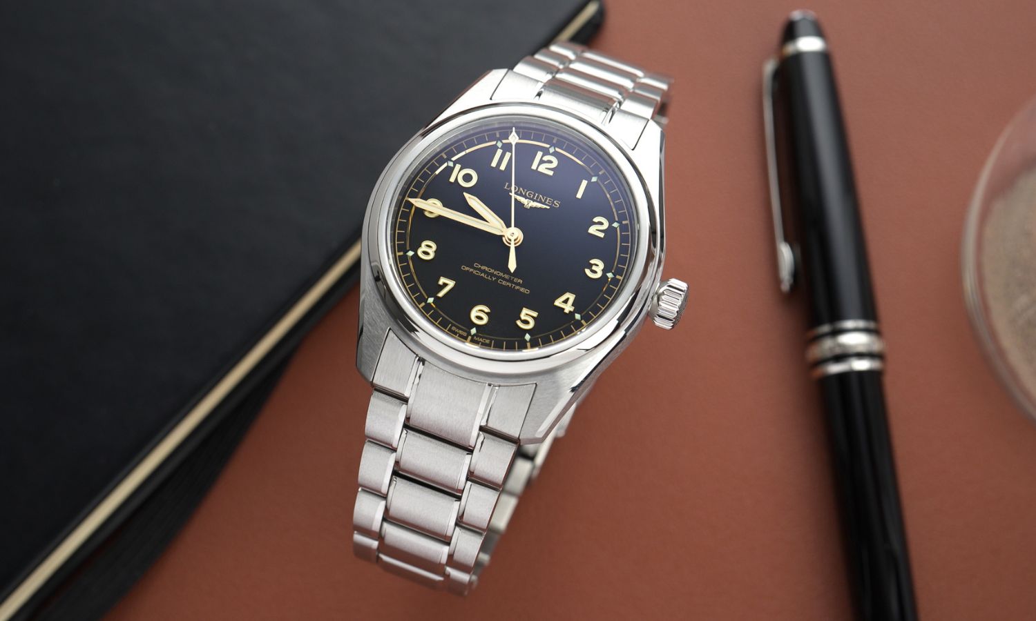 Longines Spirit Pilot Referenz L3.809.4.53.6 mit schwarzem Zifferblatt am Edelstahlarmband von vorne abgebildet stehend neben einem schwarzen Kugelschreiber vor braunem Hintergrund
