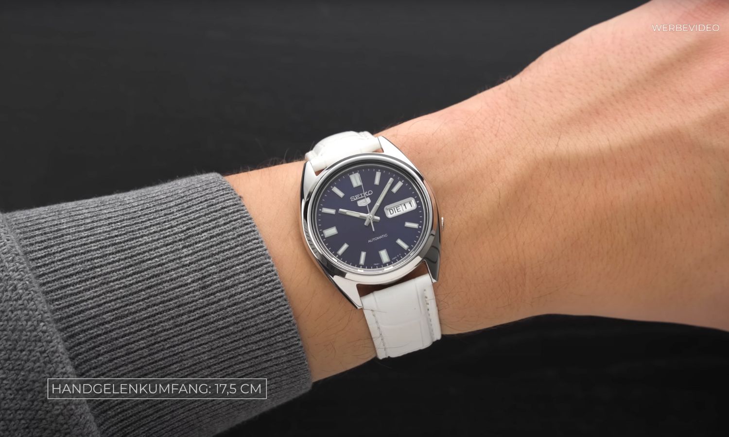 Seiko 5 SNKD97 mit dunkelblauem Zifferblatt am weißen Lederarmband an einem Handgelenk mit dunkelgrauem Ärmel getragen vor schwarzem Hintergrund