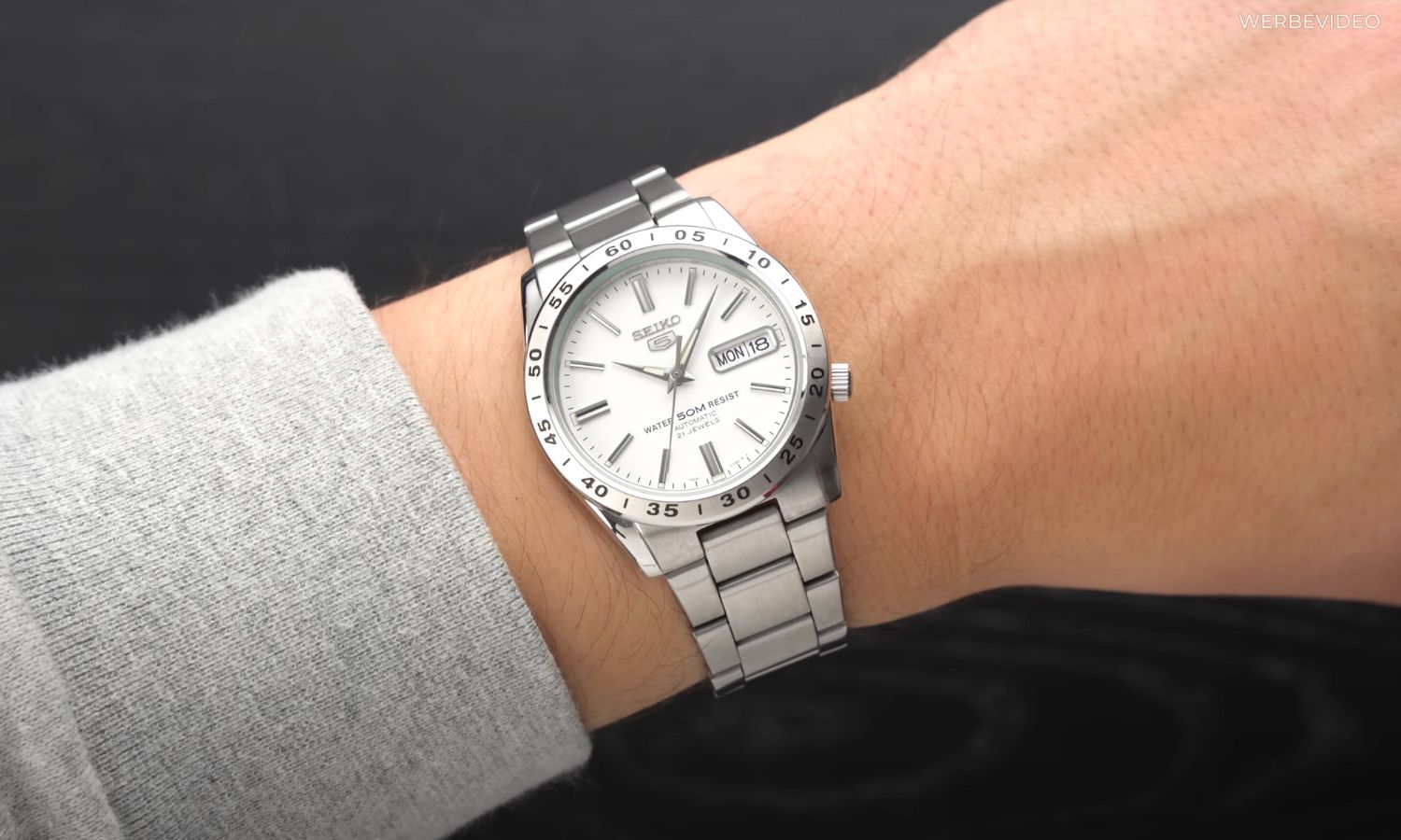 Seiko 5 SNXS77 mit weißem Zifferblatt am Edelstahlarmband an einem Handgelenk mit grauem Ärmel getragen vor schwarzem Hintergrund