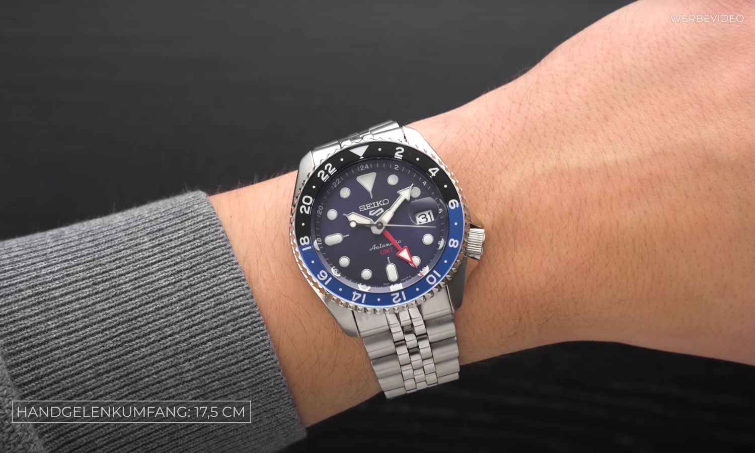 Seiko 5 Sports Automatik GMT mit dunkelblauem Zifferblatt am Edelstahlarmband an einem Handgelenk mit dunkelgrauem Ärmel getragen vor schwarzem Hintergrund