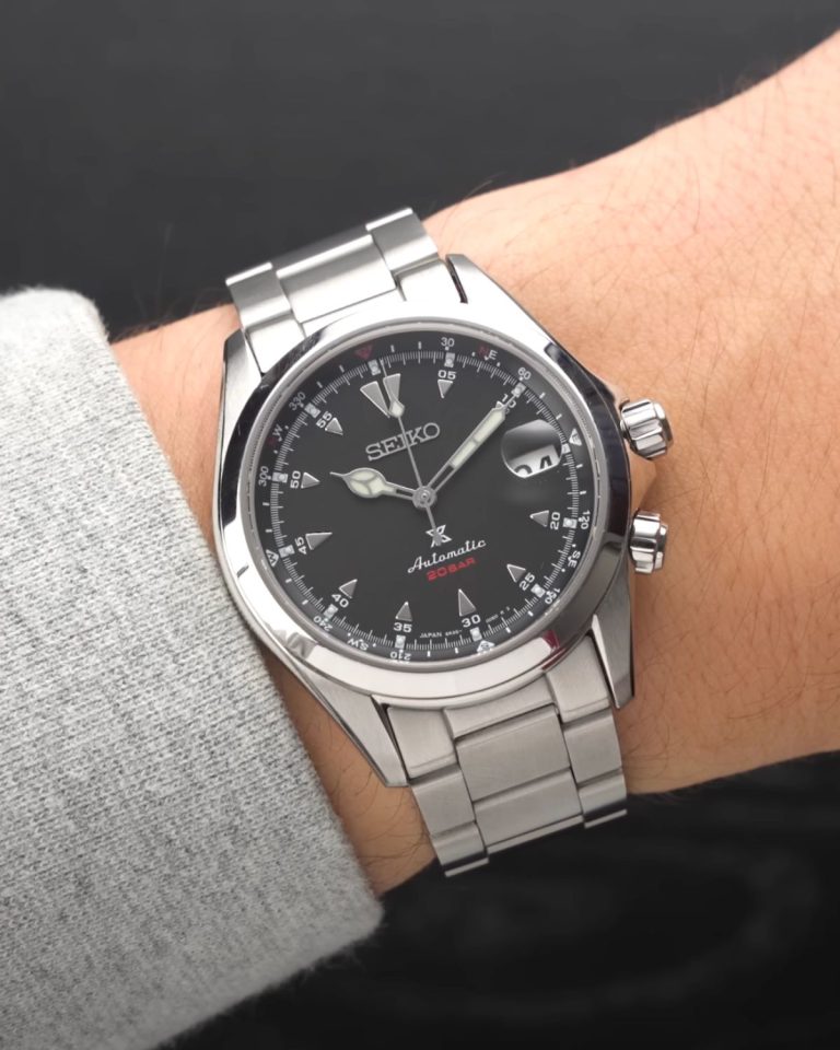 Seiko Alpinist SPB117 mit schwarzem Zifferblatt am Edelstahlarmband an einem Handgelenk mit grauem Ärmel getragen vor schwarzem Hintergrund