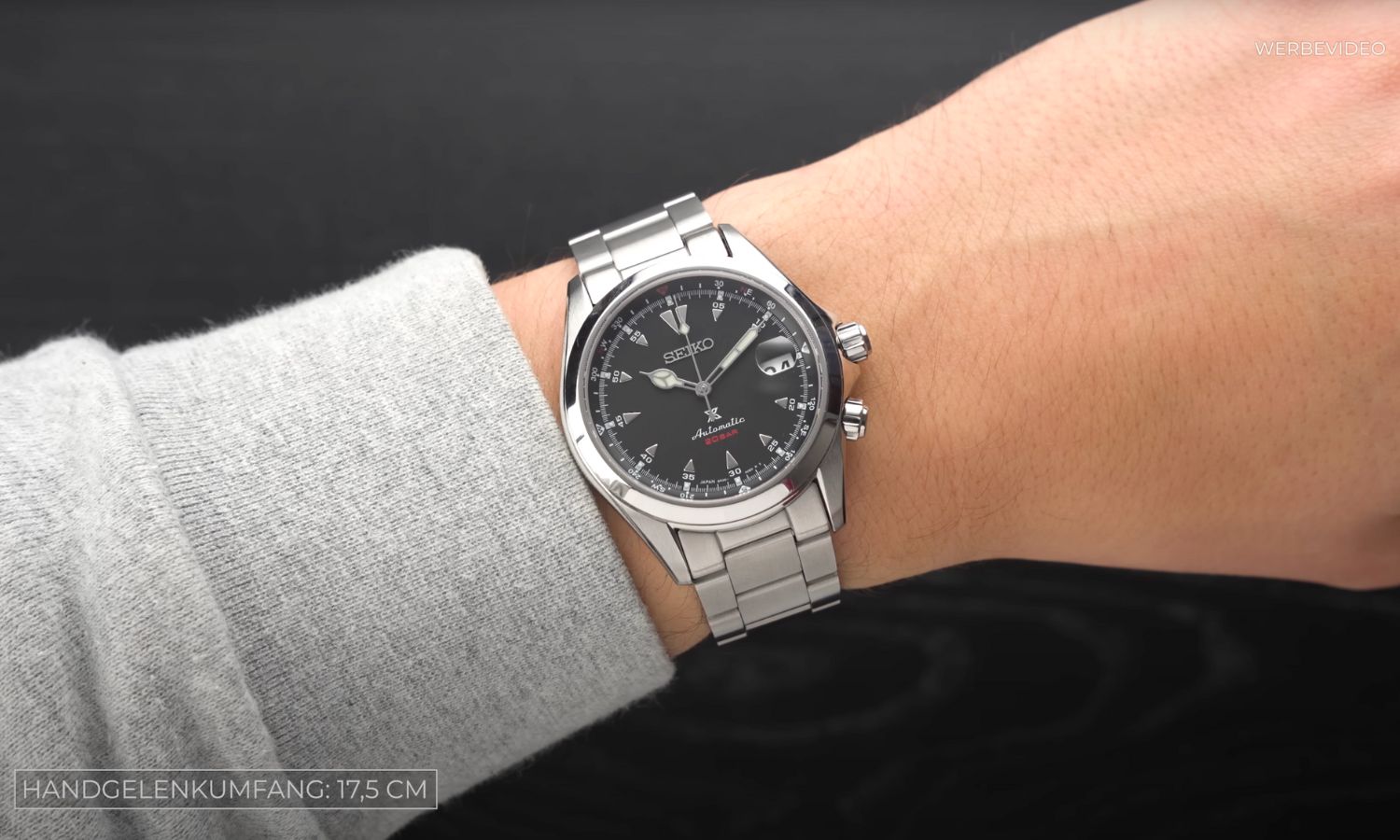Seiko Alpinist SPB117 mit schwarzem Zifferblatt am Edelstahlarmband an einem Handgelenk mit grauem Ärmel getragen vor schwarzem Hintergrund