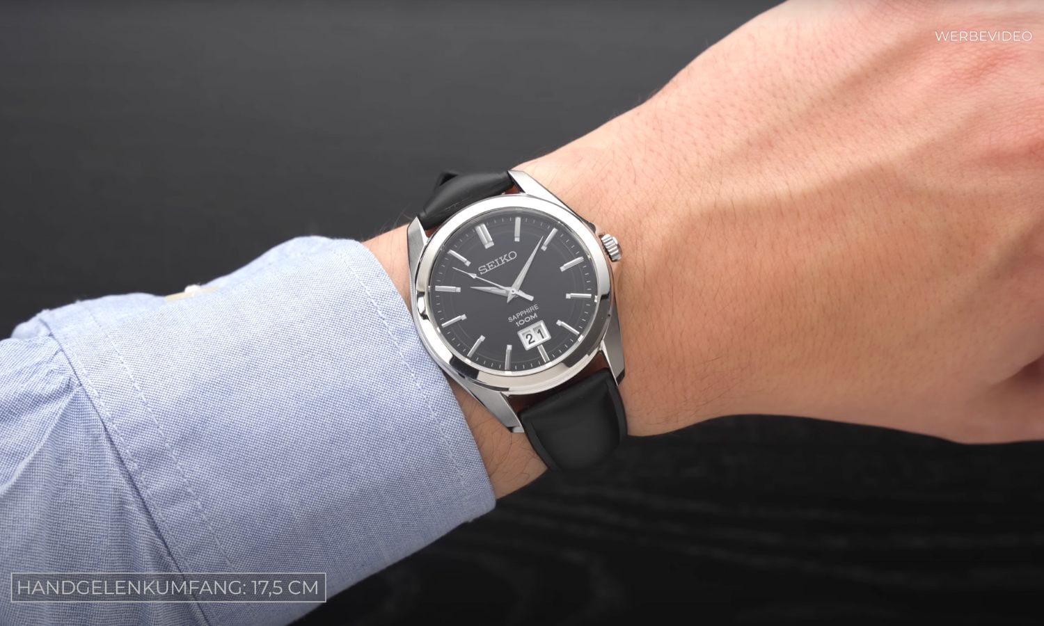 Seiko Quarz Big Date Referenz SUR009P1 mit schwarzem Zifferblatt am schwarzen Lederarmband an einem Handgelenk mit hellblauem Ärmel getragen vor schwarzem Hintergrund