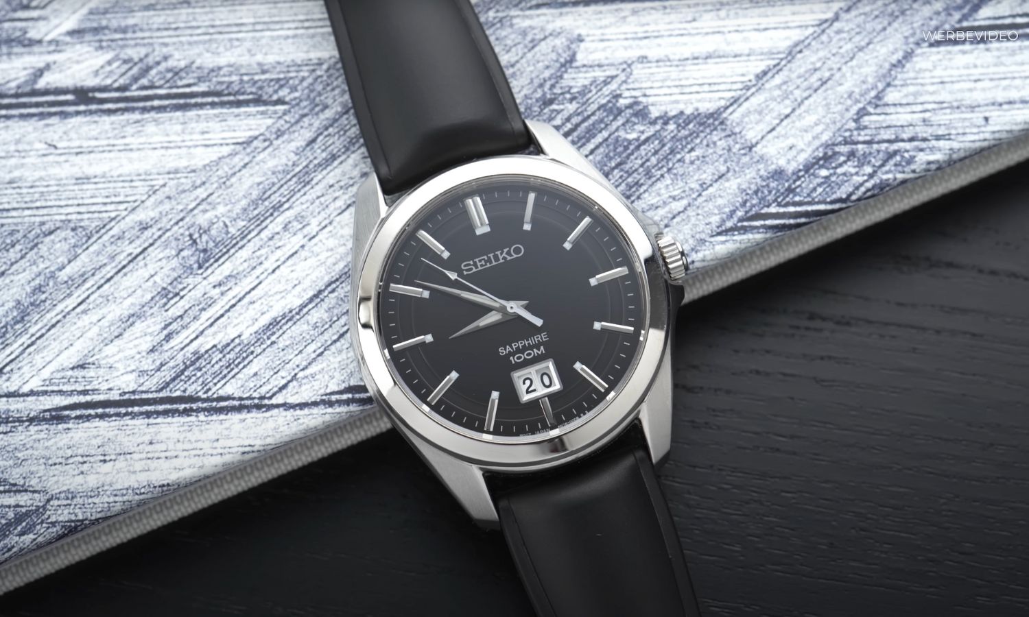 Seiko Quarz Big Date Referenz SUR009P1 mit schwarzem Zifferblatt am schwarzen Lederarmband liegend auf hellem Hintergrund
