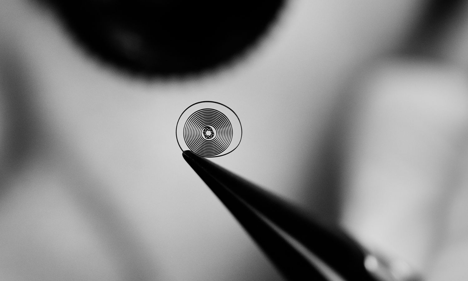 TAG HEUER TH-Carbonspring in einer Pinzette gehalten vor verschwommenem grauen Hintergrund