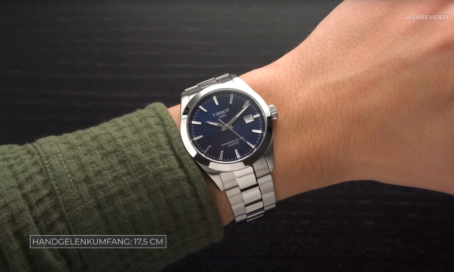 Tissot Gentleman Powermatic 80 mit dunkelblauem Zifferblatt am Edelstahlarmband an einem Handgelenk mit dunkelgrünem Ärmel getragen vor schwarzem Hintergrund