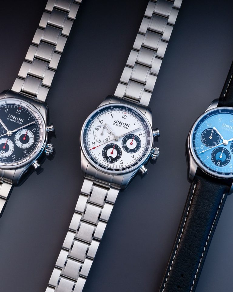 UNION Glashütte Belisar Chronograph Referenz D009.227.11.052.00 mit schwarzem Zifferblatt am Edelstahlarmband links, UNION Glashütte Belisar Chronograph Referenz D009.227.11.012.00 mit weißem Zifferblatt am Edelstahlarmband in der Mitte und UNION Glashütte Belisar Chronograph Referenz D009.227.16.042.00 mit blauem Zifferblatt am schwarzen Kalbslederarmband rechts liegend vor grauem Hintergrund