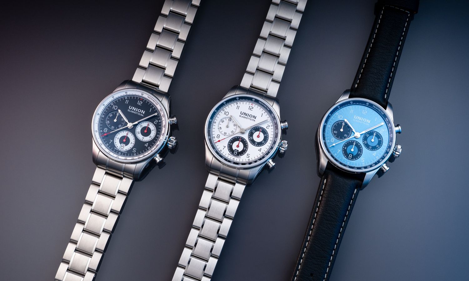 UNION Glashütte Belisar Chronograph Referenz D009.227.11.052.00 mit schwarzem Zifferblatt am Edelstahlarmband links, UNION Glashütte Belisar Chronograph Referenz D009.227.11.012.00 mit weißem Zifferblatt am Edelstahlarmband in der Mitte und UNION Glashütte Belisar Chronograph Referenz D009.227.16.042.00 mit blauem Zifferblatt am schwarzen Kalbslederarmband rechts liegend vor grauem Hintergrund